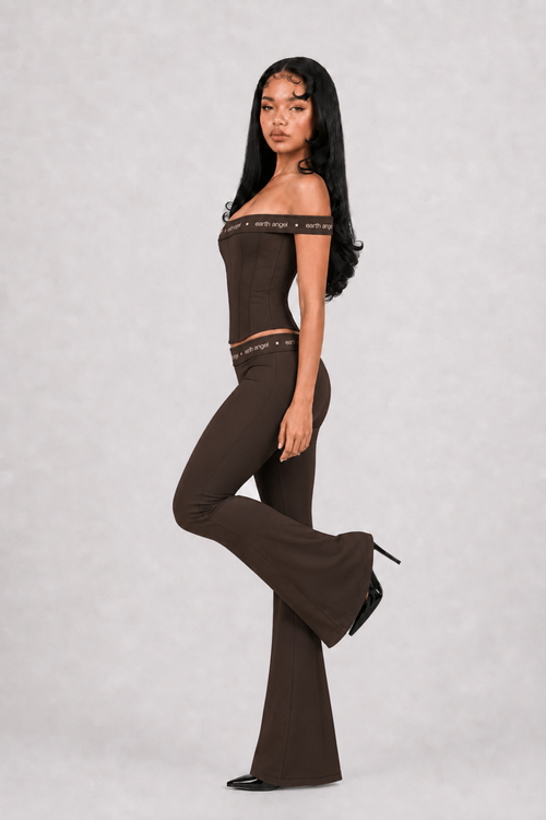 Woman wearing a brown off-shoulder top and flared pants on a light gray background
